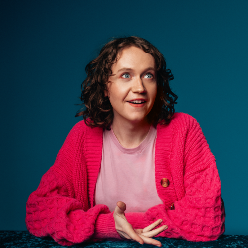 Dee sat at a table mid conversation wearing a pink shirt and cardigan..