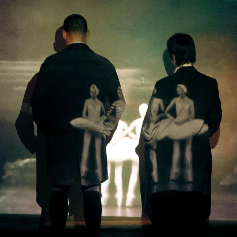 Two figures in suits stand with the faces to a wall. There are layers of photos on top including one of a ballet troupe.