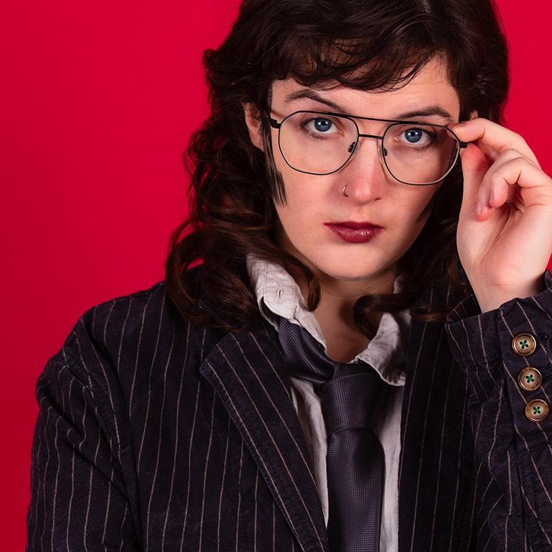 A photo Roger wearing a pinstripe suit and loose tie with sideburns and glasses.