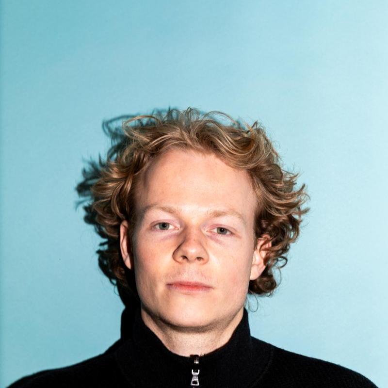 Alfie looks straight to camera, passport photo style, on a light blue backdrop wearing a black turtleneck.
