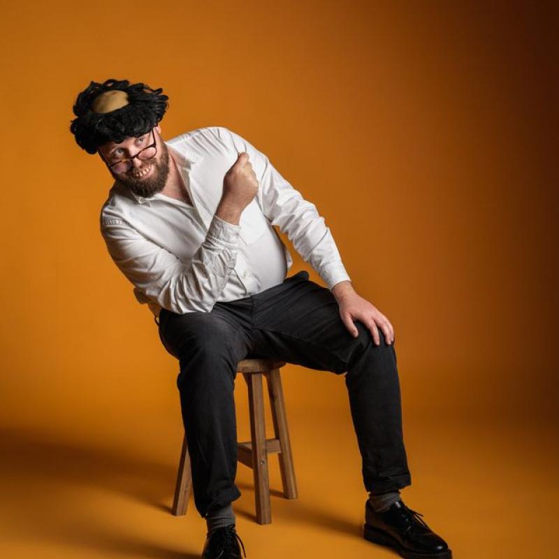 John Kearns wearing a wig and fake teeth sat on a stool.
