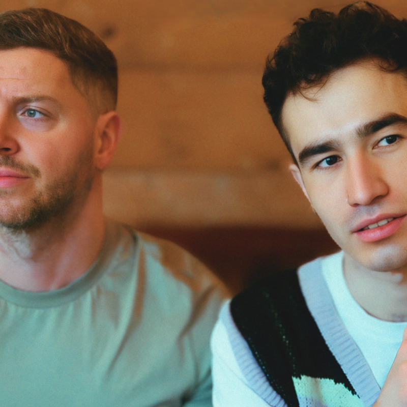 Two actors sat down with mugs in front of them both looking off camera.