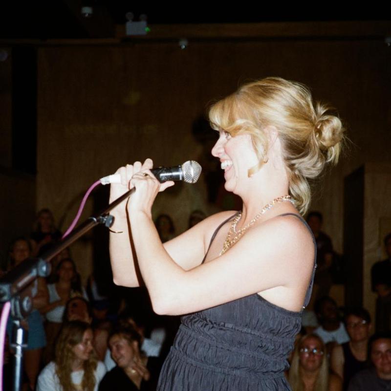 A woman with blonde hair stands on stage in front of a microphone, smiling with her hands clasped 
