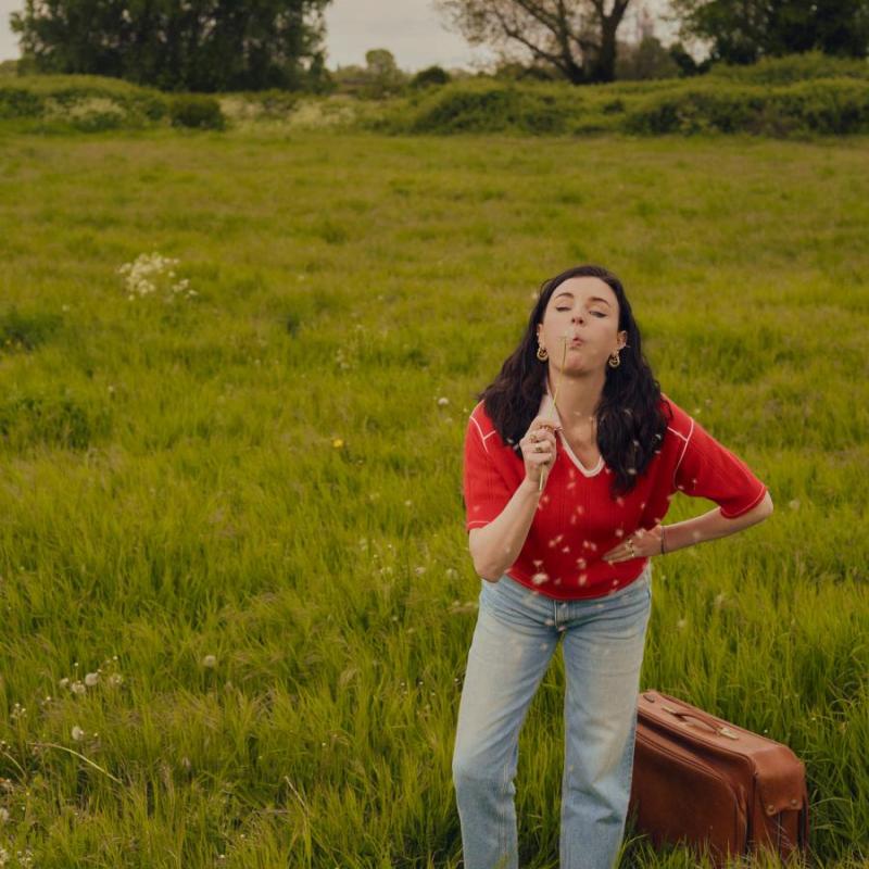 Photo of Aisling blowing a dandelion flower in a green field and stood next to a handheld brown suitcase.