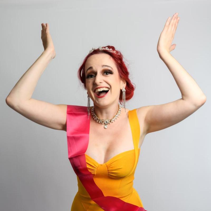 A person with red hair and elegant makeup, wearing a tiara, a bright yellow dress, pink sash, and jewelry, smiling joyfully with arms raised.