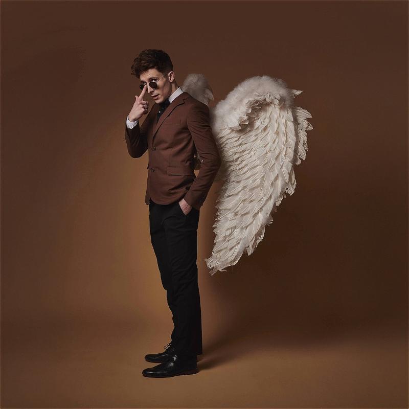 Larry is standing at a three-quarters turn to the camera, wearing angel wings, a brown business suit jacket, and round black sunglasses. He is pushing up the sunglasses with a finger. The background is brown.