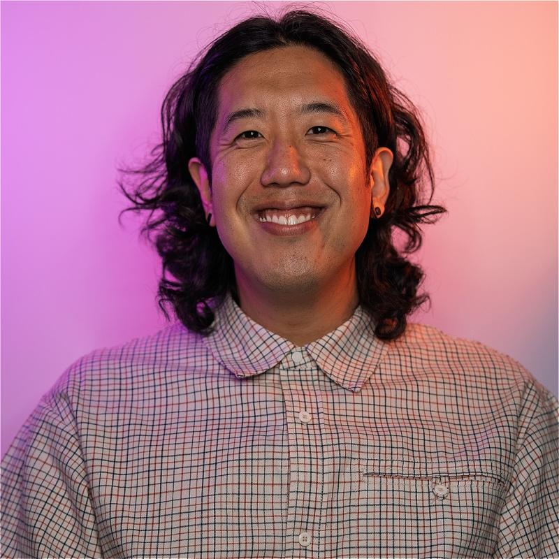A smiling Harry wearing a checkered button-down shirt stands before a pastel purple and pink background.