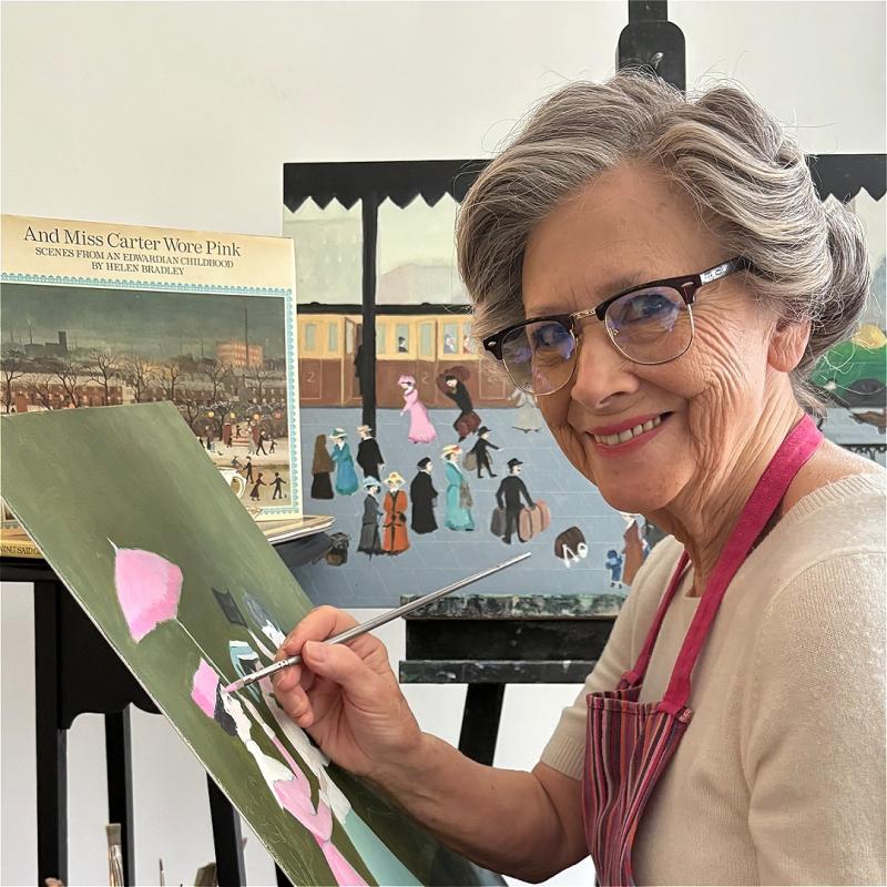 A person sits at an easel, painting, and smiles at the camera. Their grey hair is pinned up elegantly.
