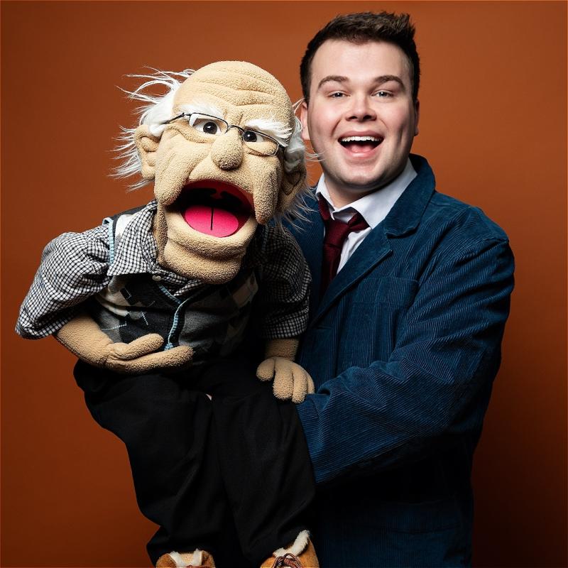 A young man holds a ventriloquist dummy of an old man before an orange background.