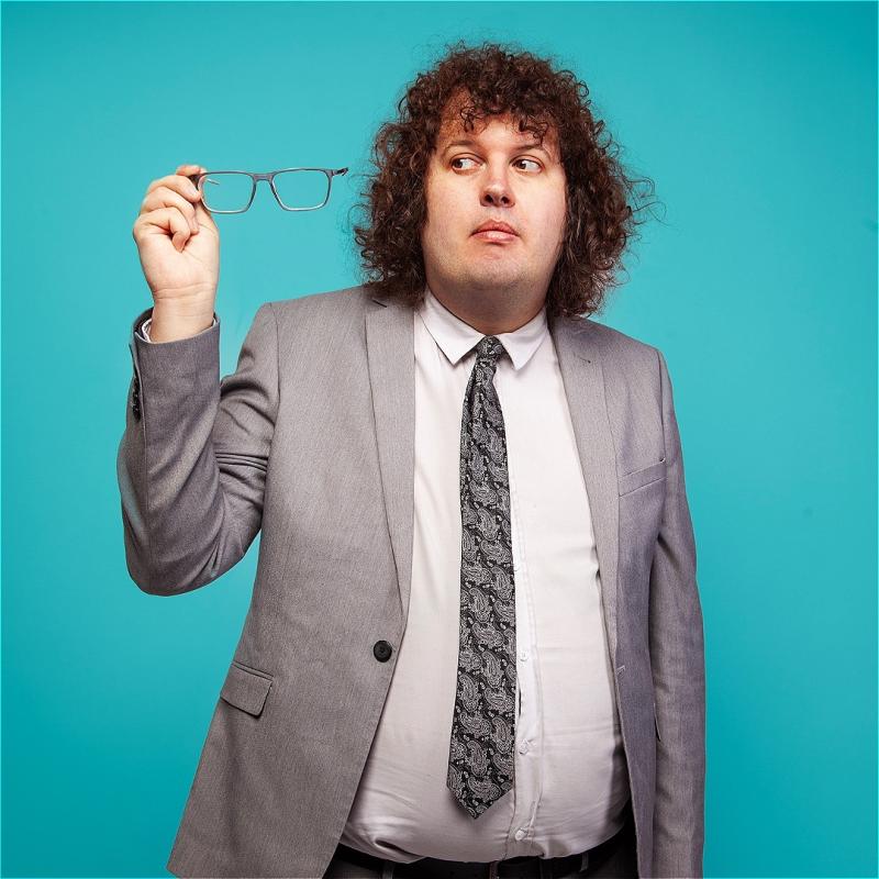 Ray is in a grey suit against a blue background, looking at their spectacles which they're holding up to the light in a gesture universally recognisable by people who use specs as 'is this still blurry'. They don't seem sure.