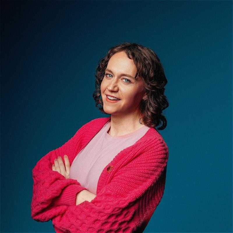 Dee is looking into the camera cheekily with their arms crossed. They are wearing a chunky knit red cardigan which contrasts against a deep blue background.
