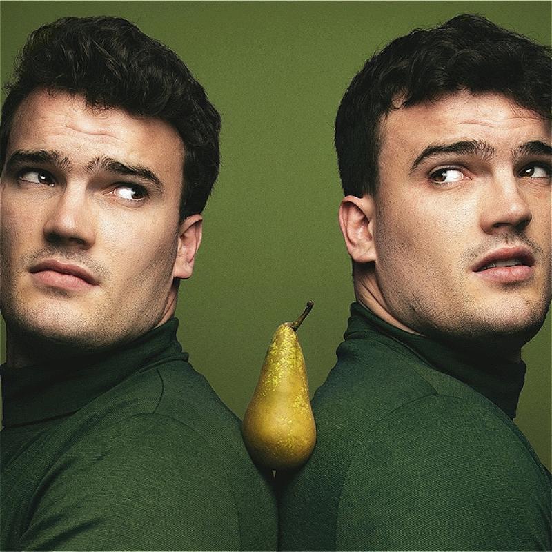 Two identical men wearing green turtleneck sweaters hold a pear between their shoulders before a green background.