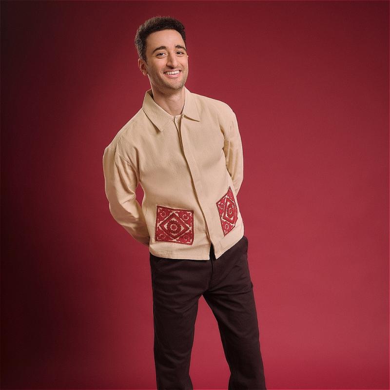 Hasan is smiling towards the camera with his hands crossed behind his back. His shirt has fabulous pockets embroidered in red patterns, matching the background.