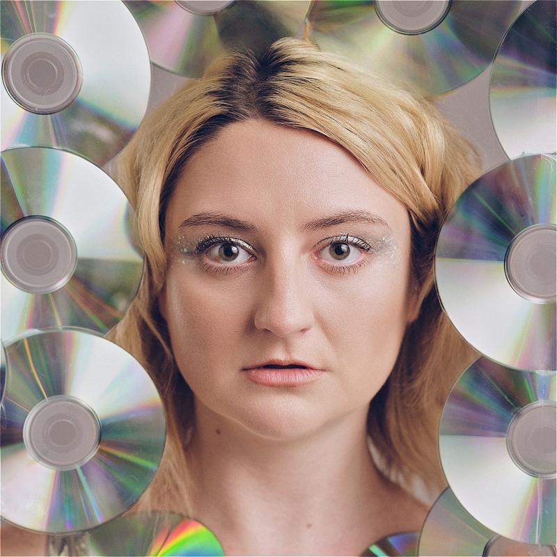 Lorna looks directly at the camera in sparkly eye shadow. They are completely surrounded by CDs, with their shiny reflective side facing us.
