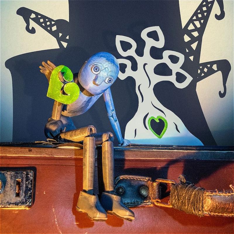A sweet little puppet with no mouth sits somewhat dejectedly on the edge of a brown leather suitcase, holding a leafy heart. There is an illustration of a small tree behind it, with branches like hearts.