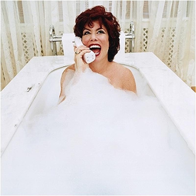 Ruby is in a luxurious, white-marble, bubble-filled bath. Bubbles cover her up to her shoulders, and she's holding an old-school white brick phone up to her ear and laughing. She's got fair skin, distinctive red hair, red lips, and a dazzling smile. 