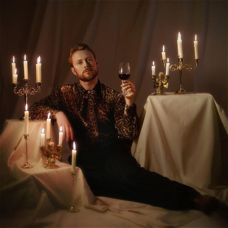 John is lying luxuriously on a draped surface, wearing leopard-print shirt and tie. They are holding up a glass of red wine and are lit by elegant white candles on multiple candlesticks and candelabras around them.