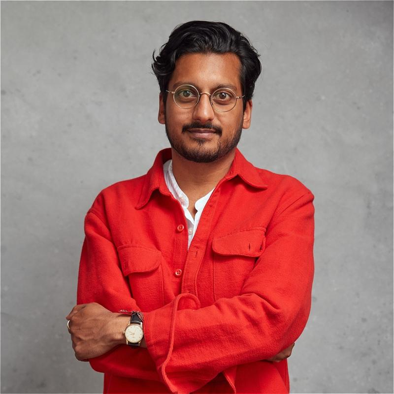 Ahir stands with their arms crossed over their chest, looking at the camera. They're wearing a bright, salmon-coloured top.