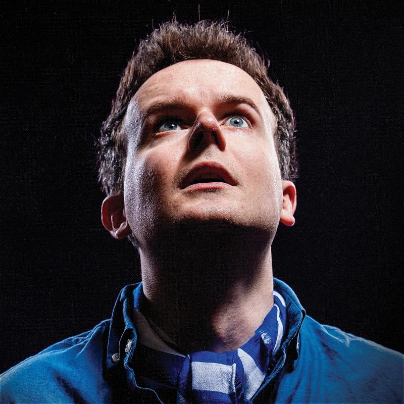 Andrew is looking up and behind the camera with a somewhat shocked look on their face. They're wearing blue against a pitch black background.