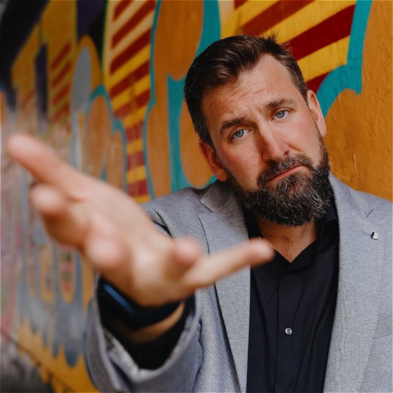 Garrett, wearing a grey suit, offers the camera his hand, before a wall with colorful paint.