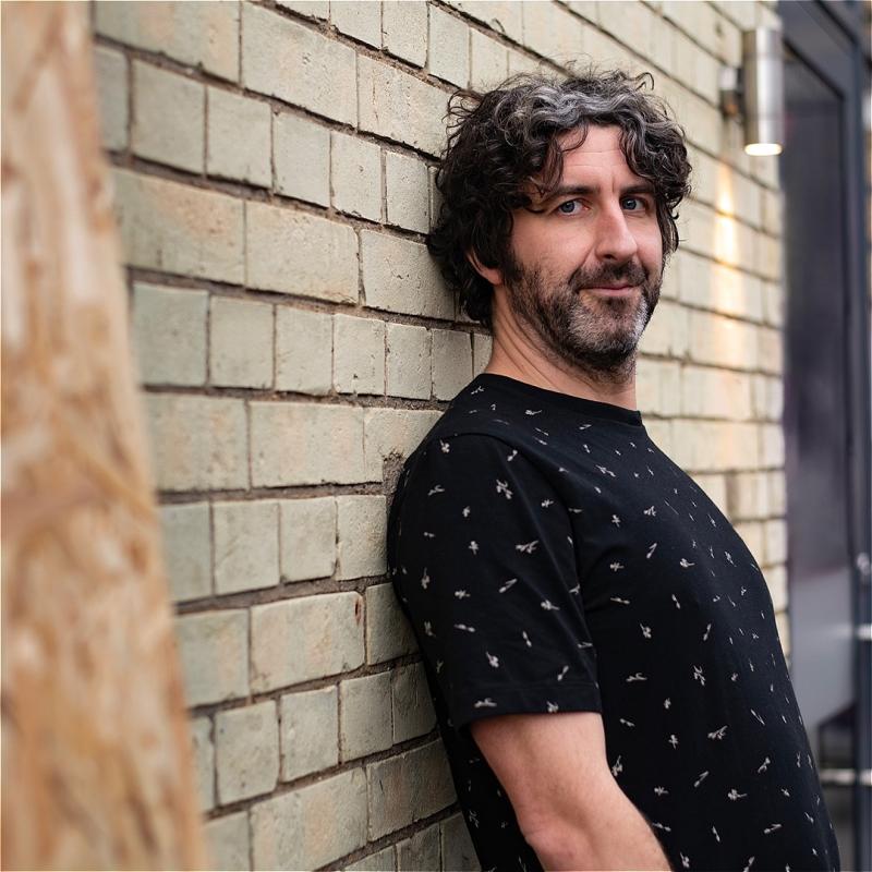 A person in a black top leans back against a brick wall and looks into the camera. They have dark curly hair, a dark beard and light skin, and they are wearing a black T-shirt. There appears to be a tree in the foreground just off to the side. 