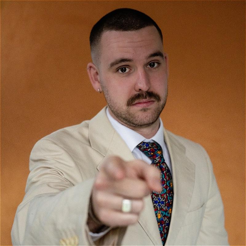 Rob is wearing a cream suit and a colourful tie against a russet background. They are pointing directly at the camera.
