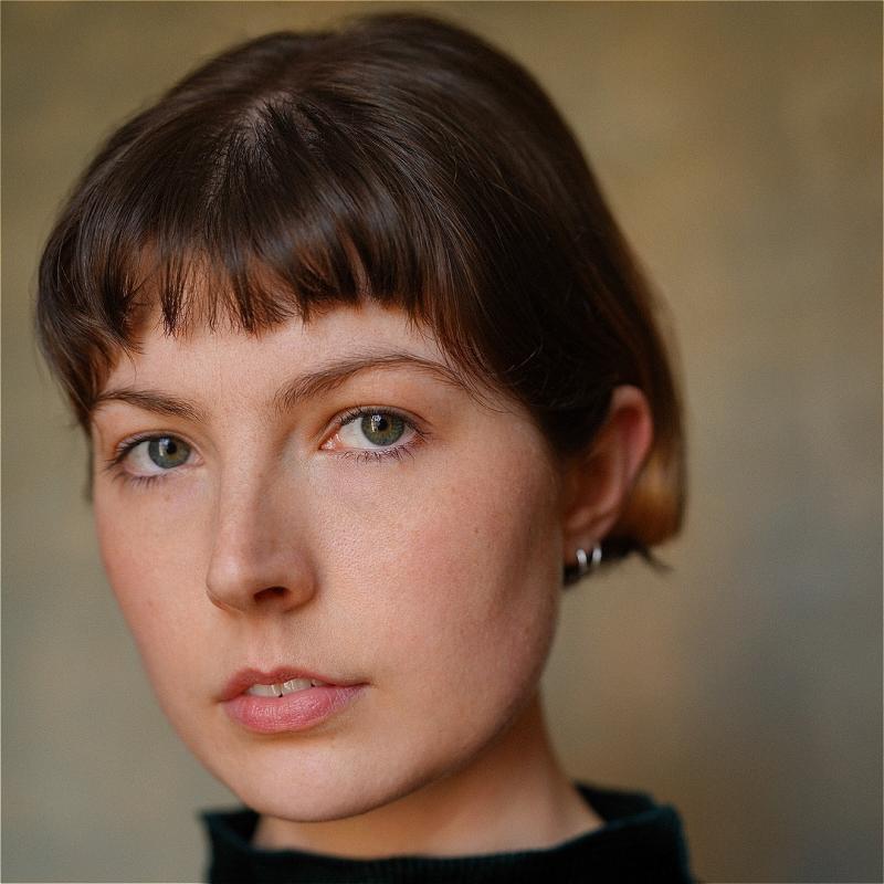 Headshot of the performer wearing a dark turtleneck sweater before a beige background.