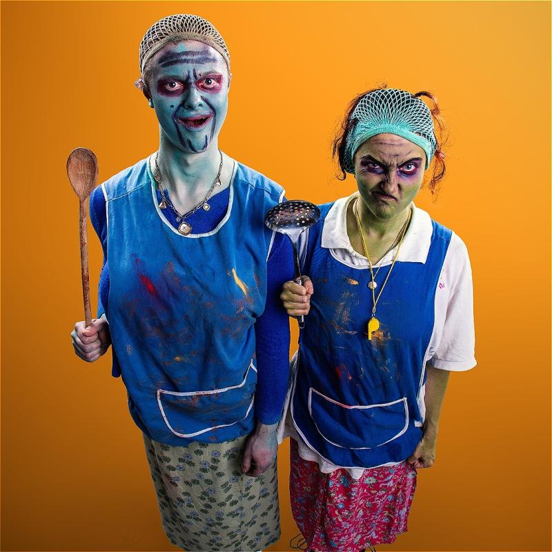 Two performers wearing blue cafeteria aprons, hair nets, and unconventional green and blue makeup hold up kitchen utensils before an orange background.