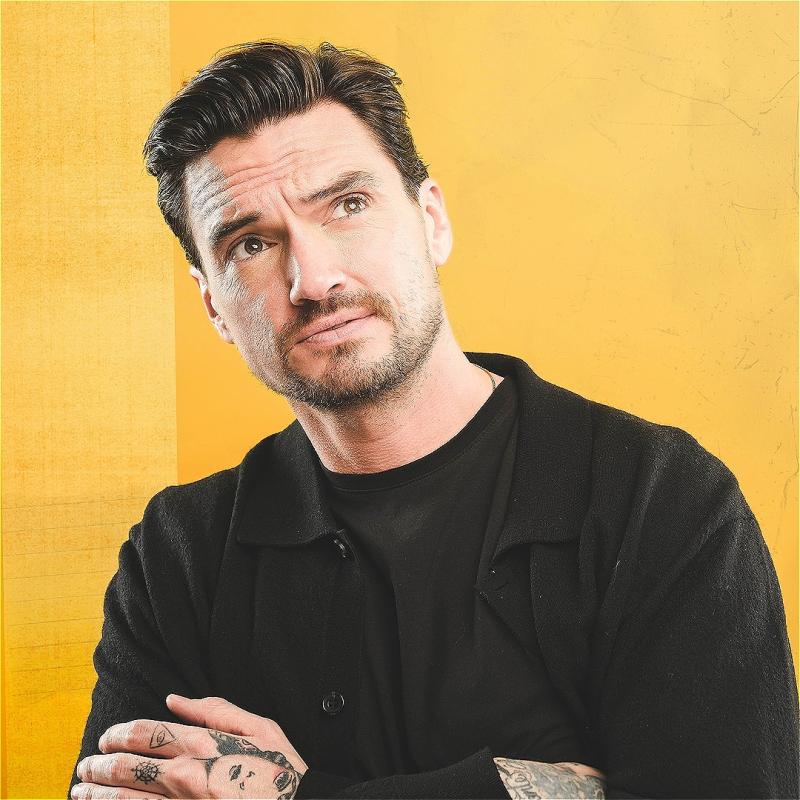 Jack is crossing their arms and looking off-camera. They are wearing black which contrasts with the bright yellow background. Their hands are tattooed - one of these tattoos appears to be a cartoon face.