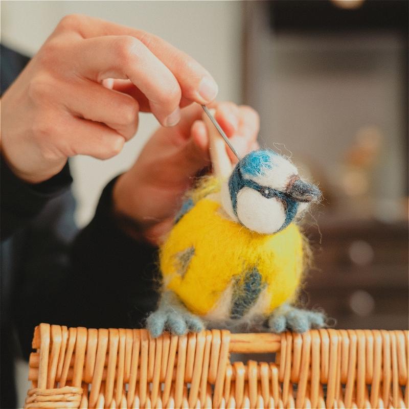 A pair of hands manipulate a blue and yellow bird made of string, on top of a wooden perch.