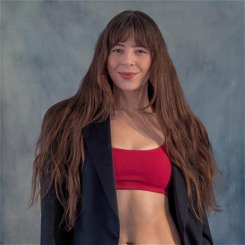 Chelsea, wearing a red sports bra over a black blazer, smiles at the camera before a grey background.