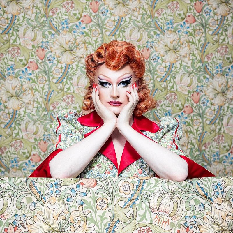 Drag queen Kate Butch stares ahead while wearing a flowery pattern dress that merges into the background, which has the same pattern.