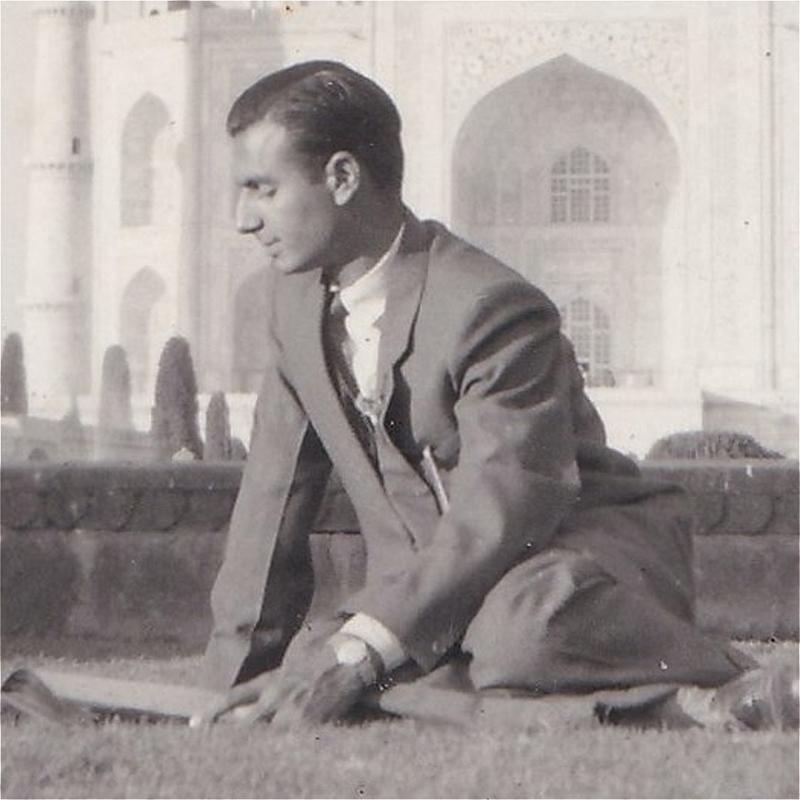 An old black and white photo of a person in a suit. Their eyes are closed and they appear to be sitting on some grass, with what looks like a fortress in the background. It has beautiful arches.