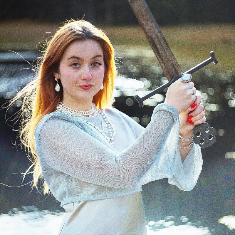 Em is wearing diaphanous white clothing and weilding a broadsword. There is a body of water in the background. They look ready to throw down.