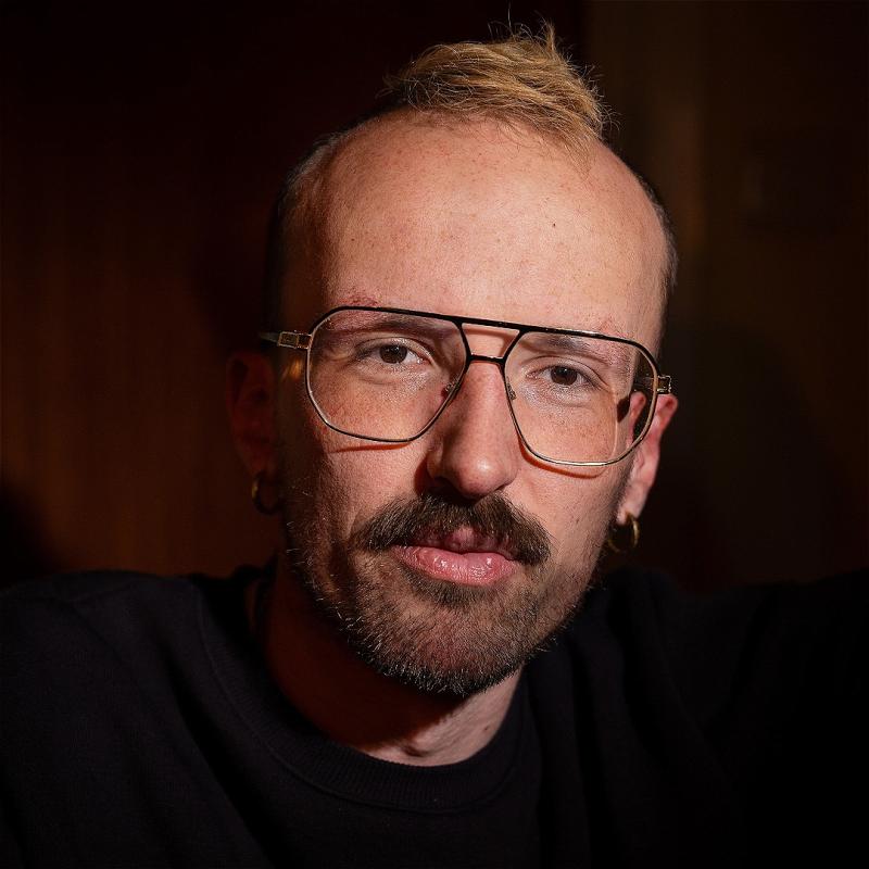 Closeup on andy, who's wearing a dark top against a dark background, and has on some fabulous aviator-style specs