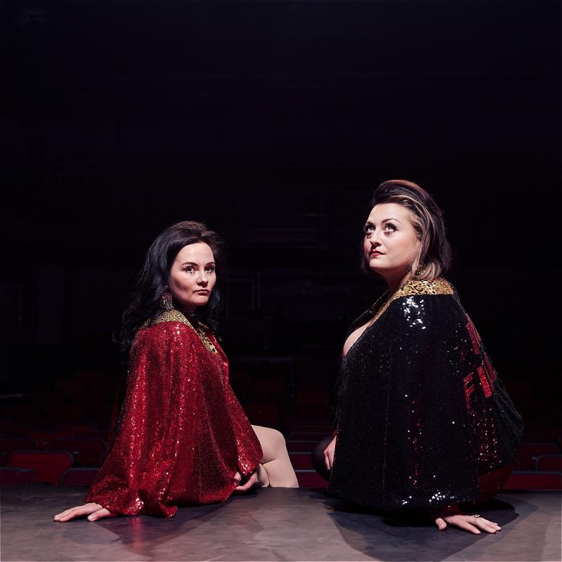Two people sitting on the edge of a stage, in a darkened auditorium. We are looking at them from behind - they're twisting back round to look at the camera. They are both wearing sparkly glittery gowns in black red and gold