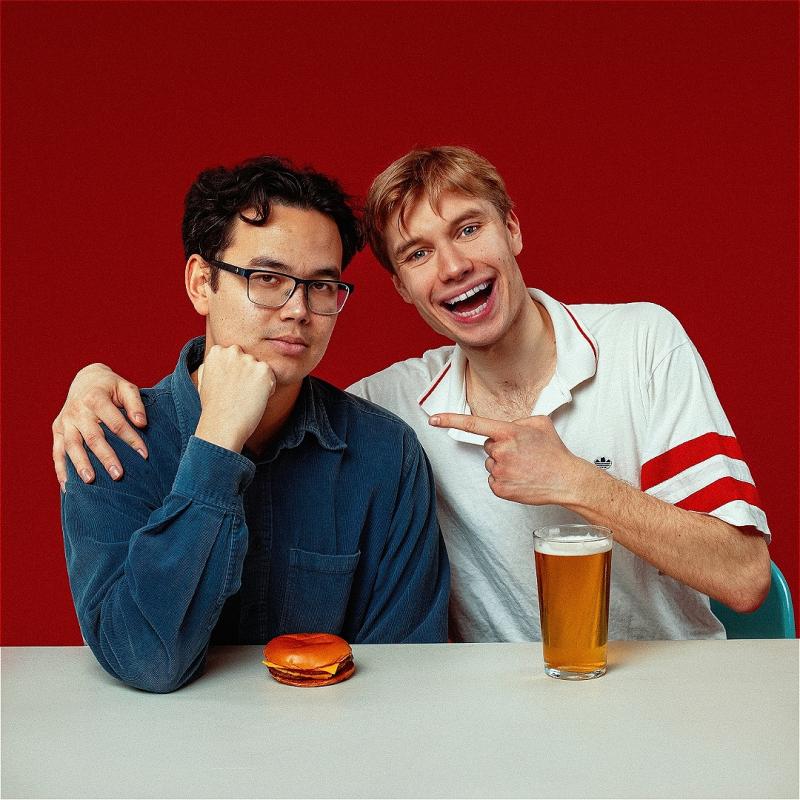 Two people sit companiably at a white surface, against a white background. One has an arm around the other, and is laughing and pointing in a friendly way as they both look to the camera. In front of them, you guessed it: one burger, and one pint. Both these items look a little disappointing in a comedic way.
