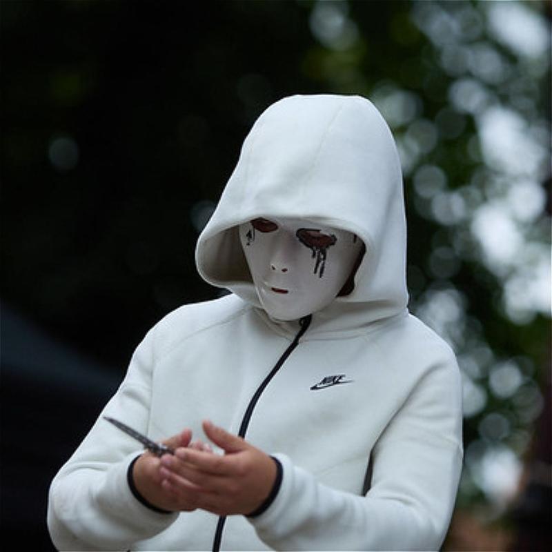 A person in a white Nike tracksuit stands with their hood up. Their face is completely covered by a mask which has black tears painted underneath the eyes. They appear to be holding something - possibly a knife