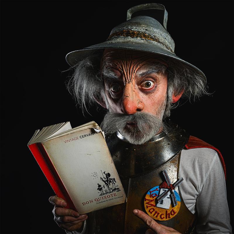 An old man stares ahead while wearing a metal breast plate, a metal helmet, and holding a tattered Don Quixote book, before a black background.