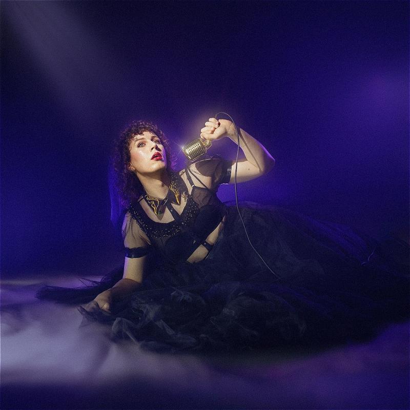 Sam lies on the floor in a fabulous black tulle dress, holding a mic up as the spotlight shines down.