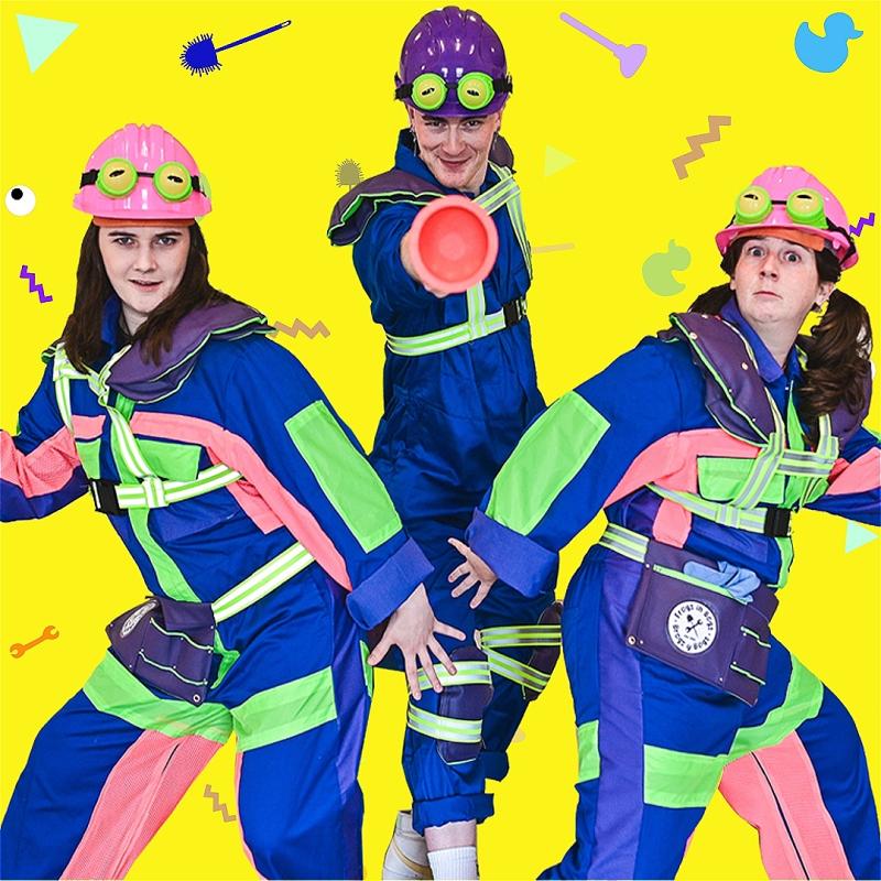 Three people wearing colorful plumber outfits  and colorful helmets with frog eyes stand before a bright yellow background.