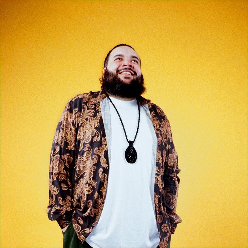 Photo of beaming Raj Poojara wearing a white shirt, a patterned overshirt, and a black necklace before a yellow background.