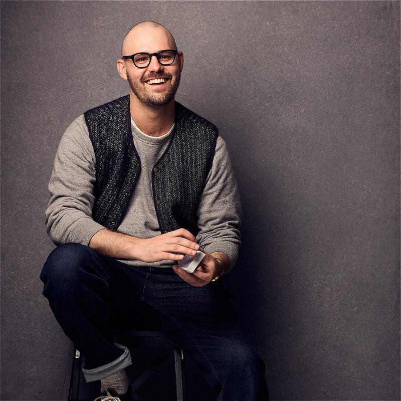 A smiling Andrew sits on a stool wearing a grey vest, jeans, and holding a deck of cards, before a grey background.