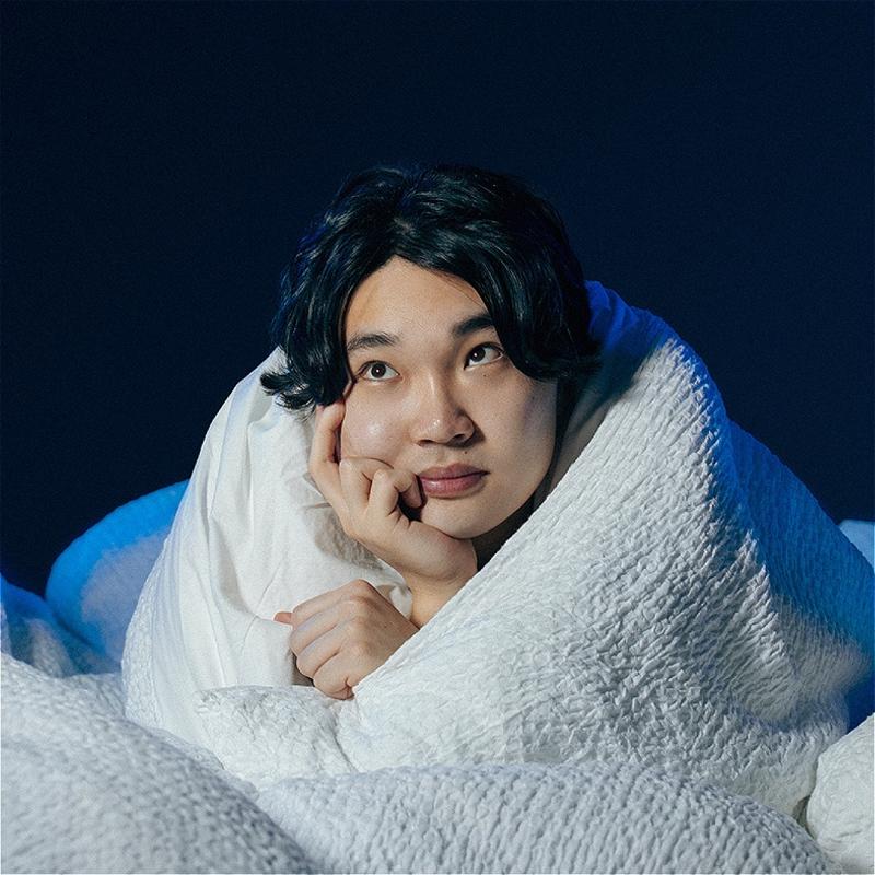 Li Jin Hao is snugly wrapped in a thick duvet, looking ponderously past the camera with their chin resting on their hand, which is all that extends from the duvet..