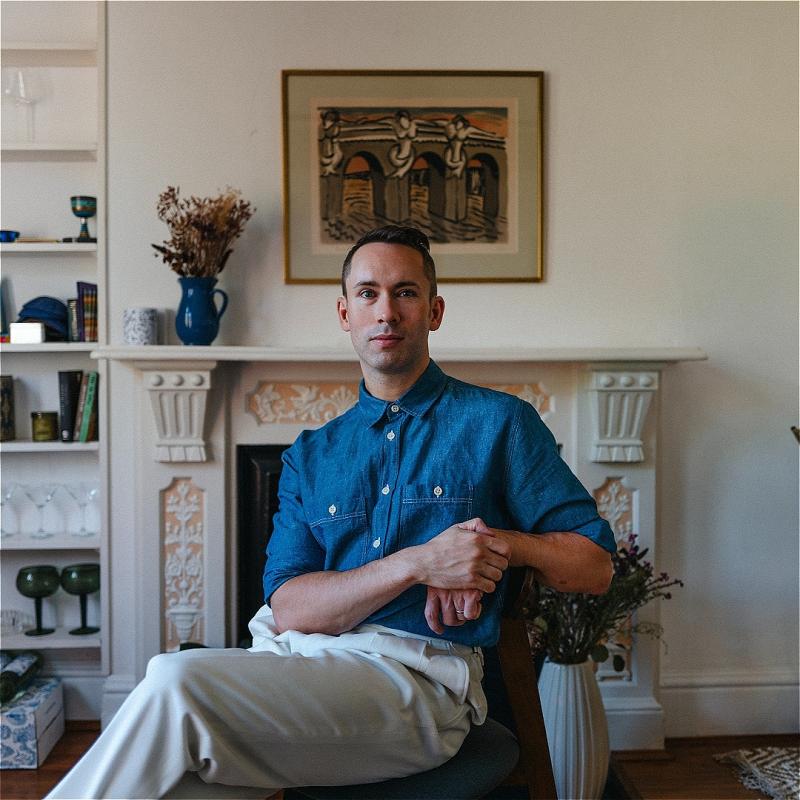 A man wearing a blue button-down shirt and white pants sits before a white living room with a fireplace, a painting and a blue vase over it.