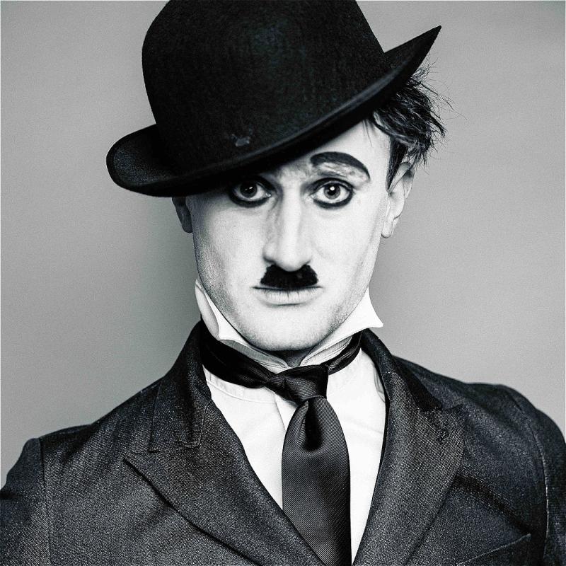A black and white image of the performer dressed as Charlie Chaplin with a bowl hat, jacket, and tie, before a grey background.