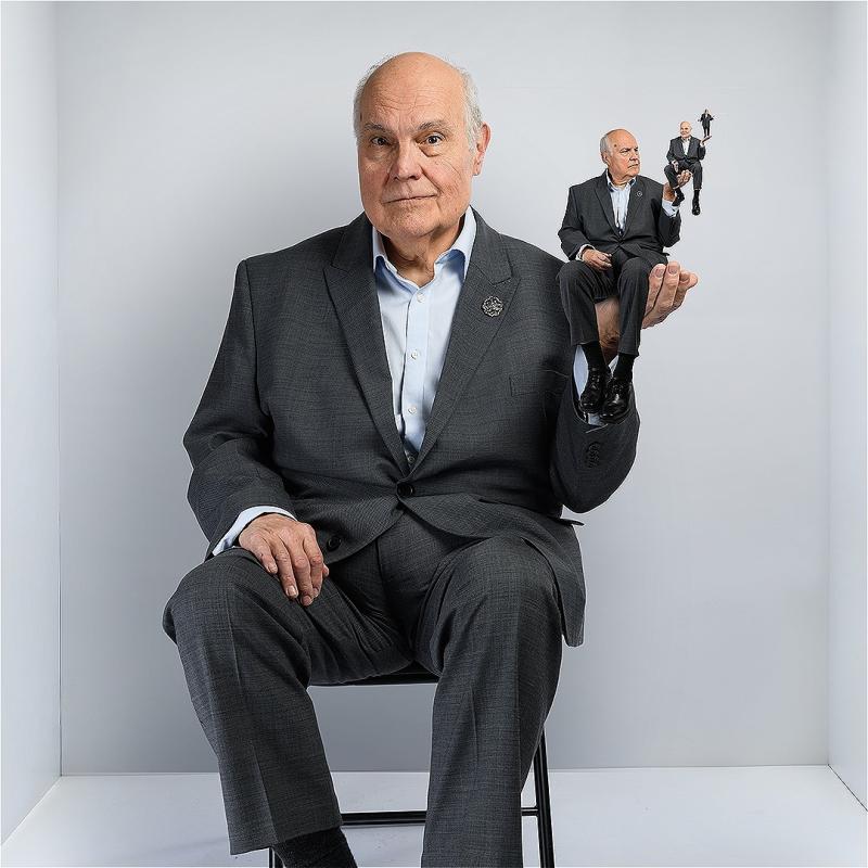 Mike sits in a chair in a small white box of a room. He's holding up a hand where a miniature of them sits, who in turn is holding up a hand that's holding another still smaller miniature twin, and so one for at least two more iterations. 