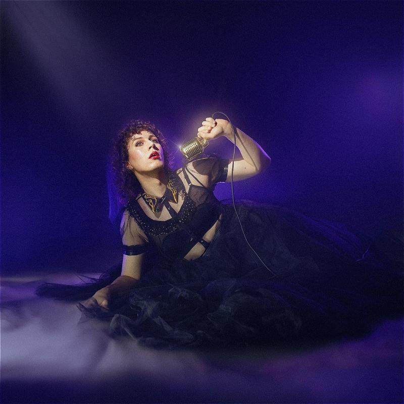 Sam in a sheer black dress lounging on the floor holding a vintage microphone. There is a purple smoky filter over the image.