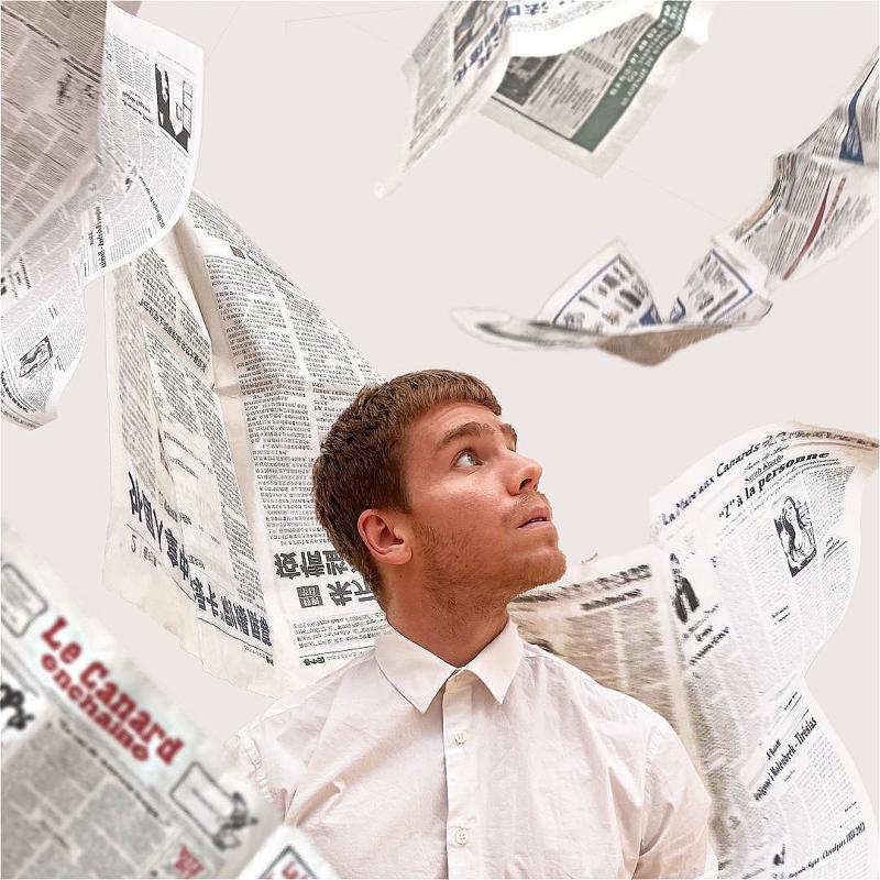 A person in a white button-up looks up as newspapers flutter down all around them. The background is white and it all feels very crisp.