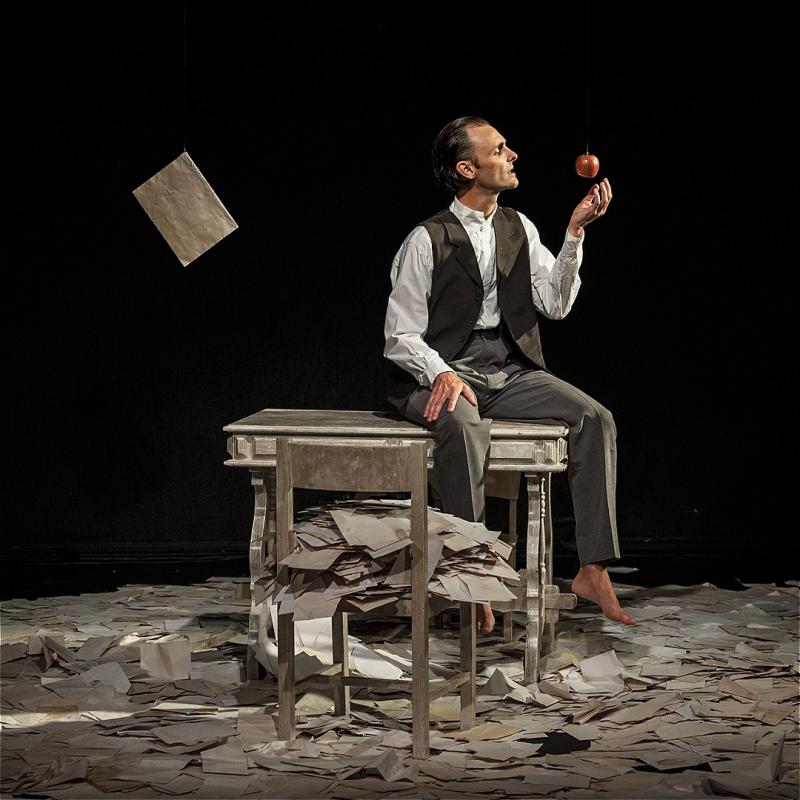 A man throws an apple as he sits on a table surrounded by countless pieces of paper, before a black background.